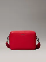 Cartera cruzada tipo 'camera bag' de Calvin Klein, color rojo vibrante, con forma rectangular y esquinas redondeadas. Presenta un acabado liso y el logo metálico de la marca en el centro frontal. Incluye herrajes metálicos plateados y una correa ajustable con detalles en tela.