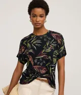 Blusa de manga corta y escote redondo, confeccionada en viscolycra con un estampado de hojas tropicales en tonos verdes, rojizos y crema sobre fondo negro.
