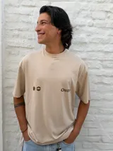 Remera oversize color beige con cuello redondo y manga corta. Presenta un estampado minimalista en color marrón en el pecho, que incluye un símbolo similar a un sol o estrella y texto.