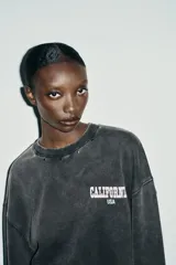 Sudadera de cuello redondo y manga larga, color gris oscuro con efecto lavado. Presenta aplicación de tachas metálicas alrededor del cuello y estampado frontal con el texto "CALIFORNIA USA" en blanco.