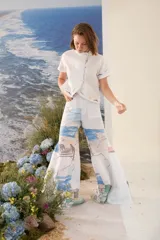 Blusa blanca de lino con mangas cortas, cuello alto con volado y cierre lateral cruzado con botones dorados. Presenta detalles bordados artesanales a contratono en el ruedo y las mangas.