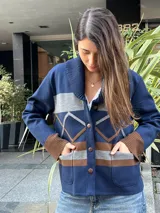 Campera de punto azul marino, con cuello smoking acanalado, bolsillos frontales aplicados y cierre de botones. Presenta bandas horizontales grises y marrones, y detalles geométricos en el frente.