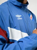 Buzo deportivo de nylon con medio cierre frontal y cuello alto. Presenta un diseño de bloques de color en azul, blanco y bordó, con el escudo del Club Nacional de Football y el logo de Umbro en el pecho.