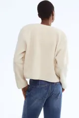 Chaqueta de sherpa suave con cuello redondo, corte holgado y cierre frontal mediante botones metálicos dorados. Presenta ribetes de cinta fina en los bordes y mangas largas.