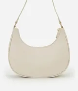 Cartera tipo baguette de diseño minimalista en color beige, confeccionada en poliuretano con acabado liso. Presenta cierre superior con cremallera, asa ajustable y detalles metálicos en tono dorado claro, incluyendo una placa con el logo de la marca en el frente.