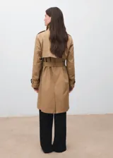 Trench coat color beige, con cuello de solapa, diseño cruzado y cierre frontal de botones. Presenta trabillas en los puños, cinturón ajustable y bolsillos laterales.