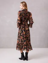 Vestido midi de gasa con estampado de arabescos en tonos cálidos sobre fondo negro. Presenta cuello alto con detalle de punto smock, mangas largas con puños elásticos y volados, y falda con tajo lateral y ruedo con volado. Incluye forro interno y cierre posterior.