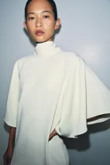 Vestido midi fluido de cuello alto y manga corta tipo capa. Presenta un diseño minimalista con bajo recto y aberturas laterales, con cierre en la espalda.