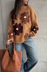 Sweater de punto grueso color camel, con diseño de flores grandes aplicadas en tonos marrones y centro crema. Presenta cuello redondo alto, mangas largas con hombros caídos y terminaciones acanaladas.