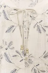 Blusa color crudo con bordados florales en azul oscuro. Presenta cuello redondo con escote pico ajustable mediante cordones con terminaciones de cuentas de madera, y mangas largas abullonadas con puños elásticos.