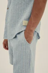 Camisa celeste a rayas verticales, de manga corta, con cuello tipo camp-collar, botones de madera y parche con logo.