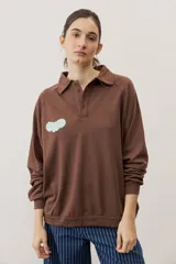 Buzo celeste de manga larga con cuello polo y cierre frontal de botones. Presenta un parche marrón bordado en el pecho.