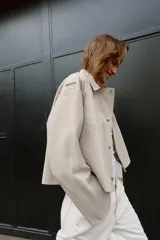 Trench corta de color beige, con cuello solapa, mangas largas con trabillas en los hombros, bolsillos delanteros de vivo y cierre frontal cruzado con botones a presión.