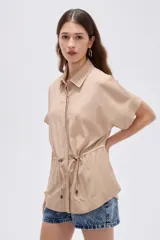 Camisa de viscosa color beige, con cuello camisero, mangas cortas, cierre frontal con botones ocultos y cordón ajustable en la cintura.