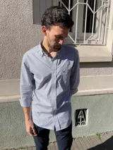 Camisa de corte clásico confeccionada en algodón, con diseño de rayas verticales finas en color rosa sobre fondo blanco. Presenta cuello abotonado, cierre frontal con botones, mangas largas con puños abotonados y un bolsillo en el pecho con el logo de la marca bordado.