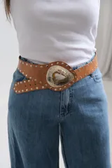 Cinto tipo faja de cuero color beige con tachas metálicas plateadas a lo largo de toda la pieza. Presenta una hebilla central grande de metal plateado con diseño grabado y una piedra natural incrustada en el centro.