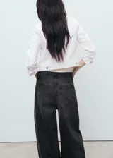Chaqueta crop estilo denim de color blanco, confeccionada en algodón con elasticidad. Cuenta con cuello camisero, manga larga con puños abotonados, cierre frontal con botones y cuatro bolsillos (dos laterales y dos de parche con solapa en el pecho).
