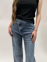 Jean de denim azul con diseño cruzado en la cintura, cierre de botón metálico y aplicaciones de strass en toda la prenda.