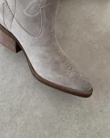 Bota corta de gamuza color marrón topo, estilo western, con bordados decorativos en hilo blanco en caña y empeine. Posee taco cuadrado de madera y punta ligeramente afilada.