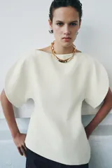 Top color crema de cuello redondo y manga corta abullonada. Presenta un bajo asimétrico y cierre en la espalda con cremallera oculta en la costura.