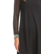 Vestido negro de seda fría con silueta evasé, cuello redondo y mangas largas de gasa transparente. Los puños son anchos y están decorados con una densa aplicación de perlas blancas.