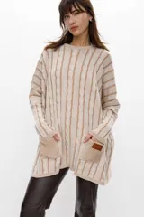 Sweater de punto color camel con diseño de trenzas verticales y acabado brillante. Presenta una silueta holgada tipo poncho, cuello redondo, mangas largas con puños ajustados y bolsillos frontales con detalle de etiqueta de cuero.