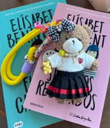 Llavero de peluche con forma de oso, vestido con una remera blanca con corazón rojo, pollera plisada negra con franjas rojas, moño a cuadros y collar de perlas. Incluye una correa de mano de cuerda amarilla con detalles trenzados.