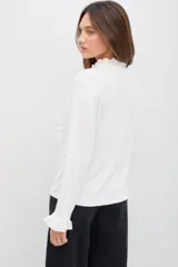 Remera de manga larga color blanco, confeccionada en tejido liviano con caída fluida. Presenta cuello alto con volados, abotonadura frontal decorativa y detalles de volados a lo largo de la abertura, además de terminaciones con volados en los puños.