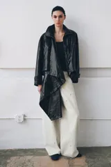 Chaqueta de efecto piel en color negro con cuello subido y bufanda desmontable integrada mediante botones. Presenta manga larga con trabillas ajustables en los puños, cierre frontal con cremallera y bolsillos laterales ocultos en las costuras. El diseño incluye un bajo con elástico en la espalda y forro interior.