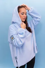 Buzo con capucha de tela de terciopelo en color celeste. Presenta un diseño holgado con aberturas laterales en el ruedo, parche con diseño de sol y texto en la manga, y detalle de logo bordado en el lateral inferior.