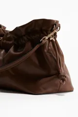 Cartera de hombro marrón de tejido revestido con diseño fruncido en la parte superior, cordón ajustable en los laterales y correa extraíble con mosquetones metálicos.