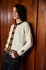 Cardigan de punto color crudo con detalles en marrón y negro. Presenta cuello redondo, cierre frontal con botones forrados y puños acanalados con rayas horizontales.