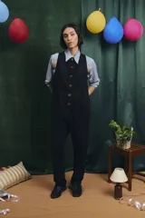 Mono largo de color negro con cierre frontal de botones vintage de distintos colores. Presenta un diseño sin mangas, con bolsillos laterales y una cinta para herramientas en la parte trasera.