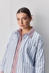 Camisa de calce relajado con diseño a rayas verticales en tonos azul y blanco, cuello clásico, mangas largas con puños abotonados y cierre frontal con botones. Presenta bolsillos aplicados en el delantero y una silueta versátil.