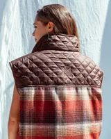 Chaleco sin mangas de diseño híbrido que combina paneles de tela acolchada capitoné en color azul marino con secciones de tejido de lana a cuadros en tonos naranja, gris y negro. Presenta cuello con forro de piel sintética marrón, cierre frontal con cremallera y ribetes en color rojo vibrante. Incluye bolsillos delanteros amplios con cierre.