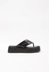 Sandalia tipo ojota con plataforma, confeccionada en cuero sintético negro. Presenta una tira ancha acolchada con costuras decorativas y una base de plataforma plana.