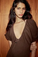 Vestido midi de color marrón, confeccionado en tejido de gasa con textura. Presenta un escote en pico con detalle de lazada frontal, mangas cortas abullonadas con terminación elástica y falda con detalle de godets que aportan volumen. Incluye forro interior a tono y cierre lateral con cremallera oculta.