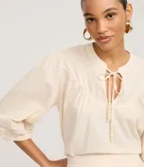 Blusa estilo bata color blanco roto, confeccionada en cambraia de algodón y lino. Presenta cuello con lazo ajustable, canesú fruncido en el busto y mangas tres cuartos con puños elásticos y bordados.