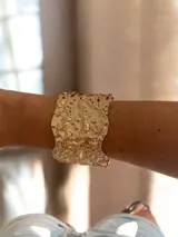 Brazalete ancho de metal dorado con diseño de superficie irregular y texturizada. Es un modelo abierto y ajustable.