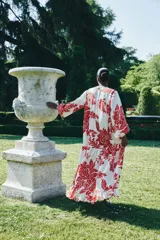 Vestido largo de tejido ligero color blanco con estampado floral rojo. Tiene cuello redondo con lazos, mangas globo largas con puños abotonados, corte holgado y falda con volantes en el ruedo.