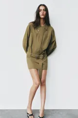 Vestido corto verde militar de cuello solapa y manga larga, con bolsillos de plastrón en el delantero y cierre frontal con cremallera oculta por sopata. Cuenta con un cinturón incorporado del mismo tejido con hebilla metálica y aberturas laterales en el bajo.