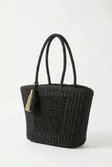 Bolso tipo tote color negro, tejido, con doble asa superior y detalle de borlas colgantes en tonos beige y negro.