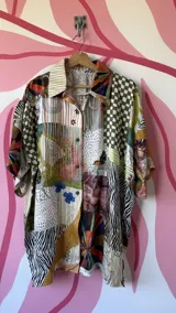 Camisa oversize de seda con estampado patchwork que combina diferentes patrones como cuadros, animal print, rayas y motivos florales. Tiene cuello camisero, botones frontales y mangas cortas.