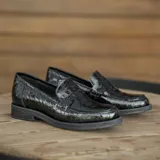 Mocasín de cuero vacuno negro con textura grabada tipo reptil. Presenta un diseño clásico con antifaz en el empeine, suela de goma en tono natural y taco bajo de madera.
