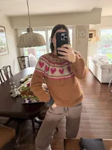 Sweater tejido a mano en mohair, de color marrón con diseño de canesú estampado con corazones fucsias y detalles geométricos en tonos beige y rosado. Presenta cuello redondo y mangas largas.