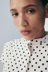 Top de popelín blanco con estampado de lunares negros, de manga corta, cuello redondo y cierre frontal con botones. Cuenta con bolsillos de parche en la parte delantera.