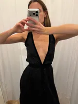 Vestido largo de seda color bronce con acabado brillante, escote profundo en V y espalda abierta. Presenta detalles en terciopelo negro que enmarcan el escote, recorren el frente y definen la cintura con una faja ajustable. Posee breteles anchos y una abertura frontal en la falda.