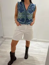 Chaleco corto en denim azul con detalle desflecado y bordado artesanal de flores en tonos celeste y verde. Estilo boho chic con escote en V y cierre frontal con botones.