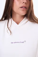 Buzo tipo canguro oversize de color blanco, con capucha y bolsillo frontal. Presenta un estampado artístico de una galaxia con estrellas en tonos violetas en la parte trasera.