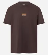 Remera de algodón peruano color marrón oscuro, corte comfort, con cuello redondo y mangas cortas. Presenta un pequeño parche rectangular estampado en el pecho con texto en tonos naranja y blanco.